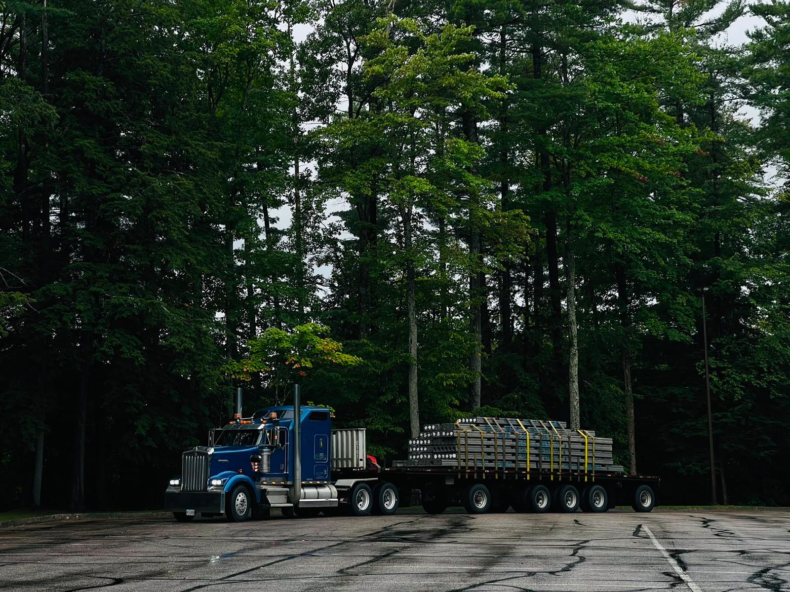 A blue Airfield Trucking Kenworth flatbed parked before a treeline in daylight